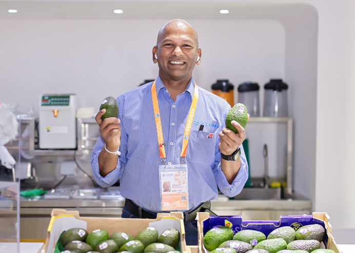 A male holding two avocados in each hand at a trade event.